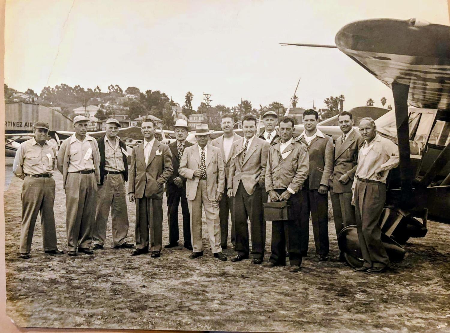 A line of men on the Martinez Airport strip beside a cabin aircraft; a hangar lettered MARTINEZ AIR; hills and houses behind