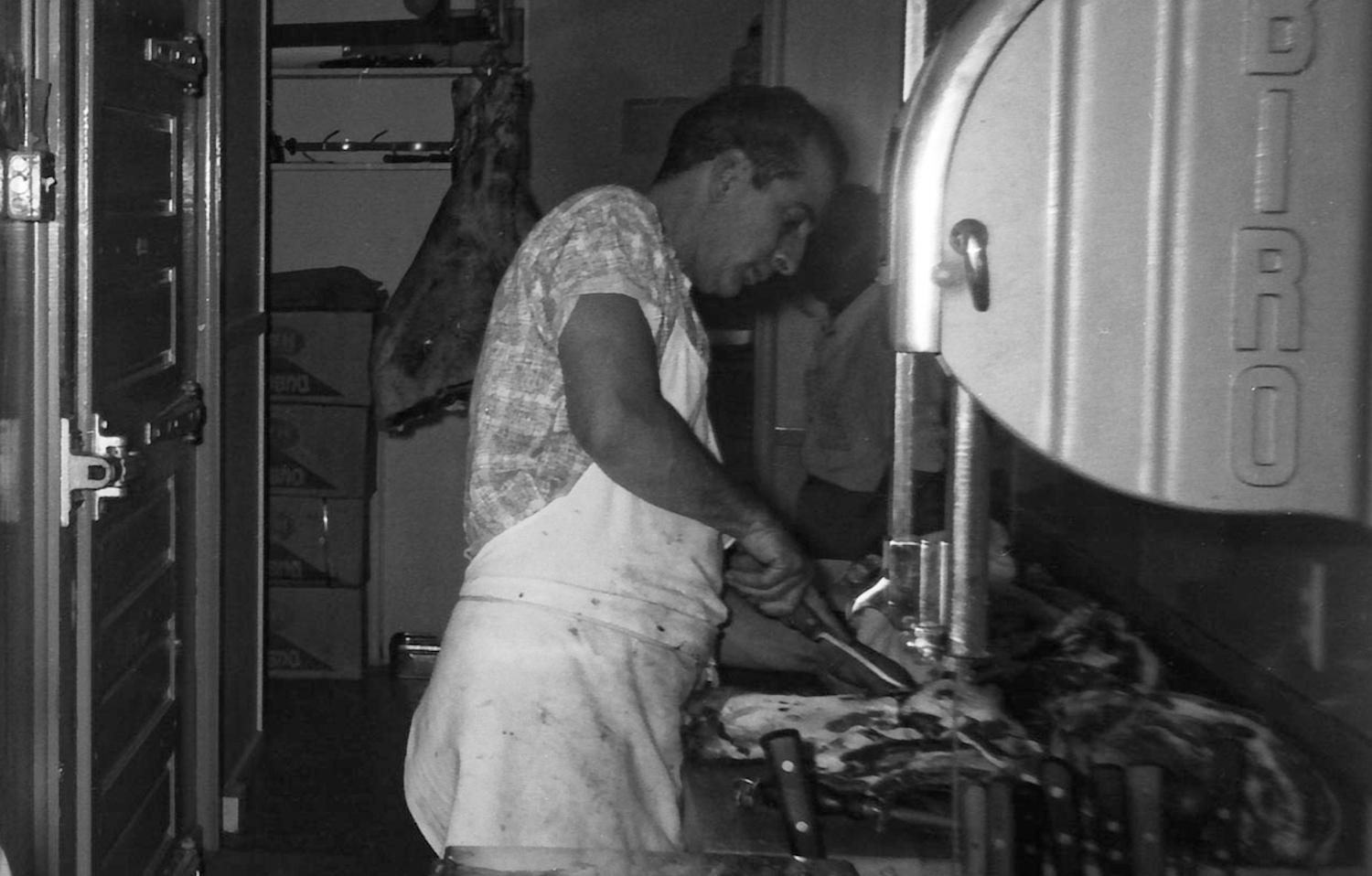 A butcher working a Biro bandsaw, a hanging carcass in the open cooler behind him, a third figure in shadow