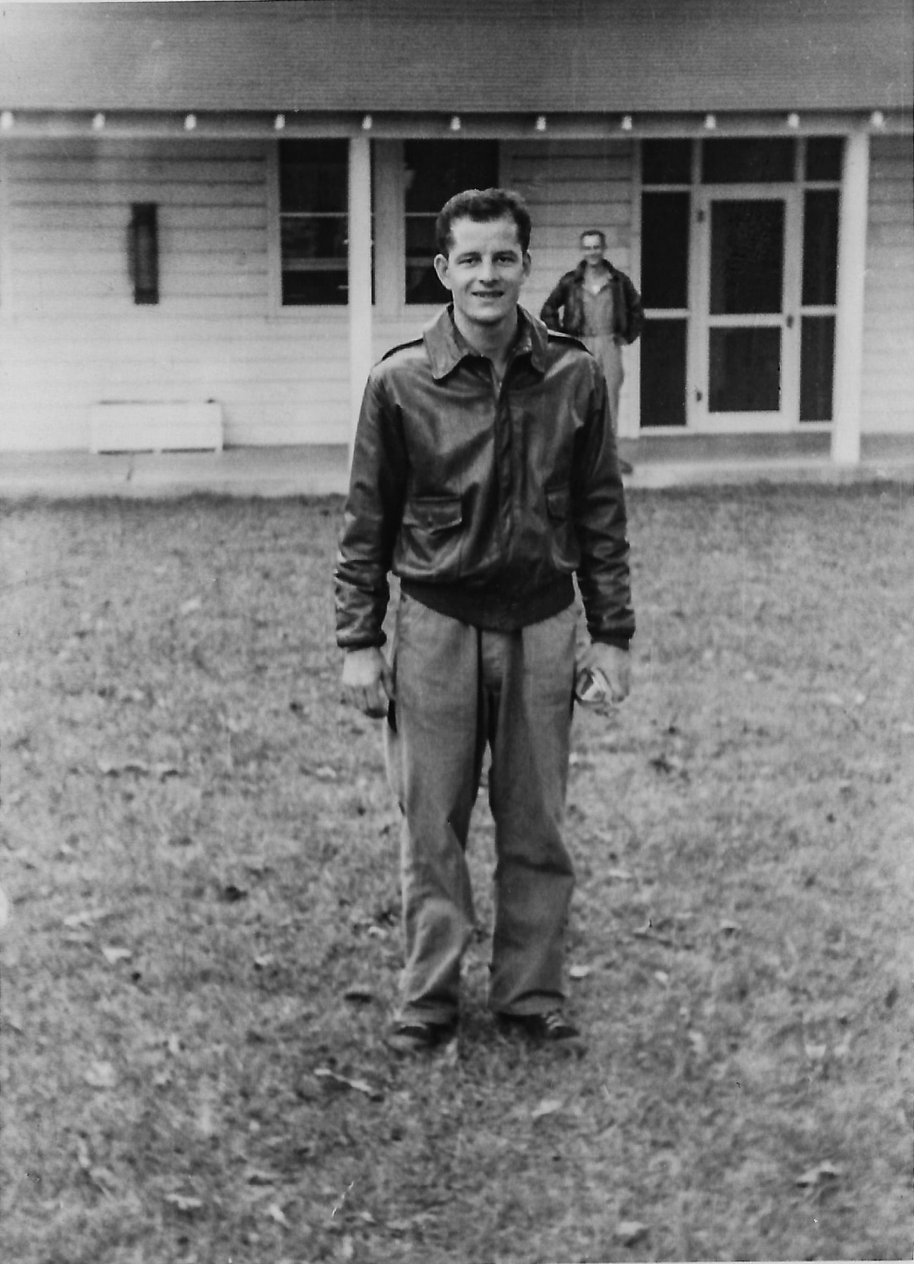 Aviation Cadet Frank Calicura standing on grass in a leather flying jacket, a man in a jacket on the barracks porch behind him