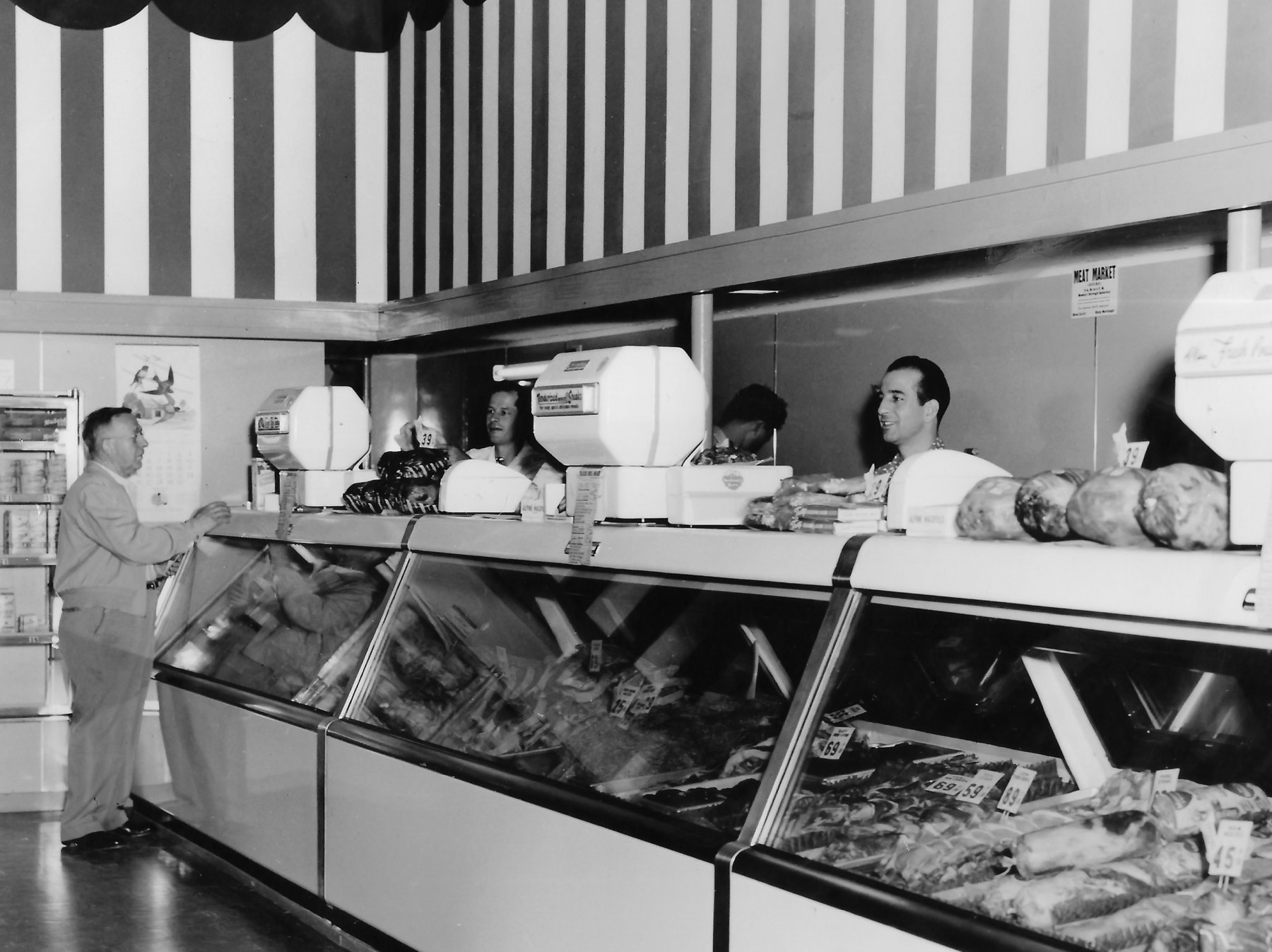 Meat market under a striped wall, a customer at left facing a long refrigerated case, men behind the counter among white scales, price cards in cents along the case