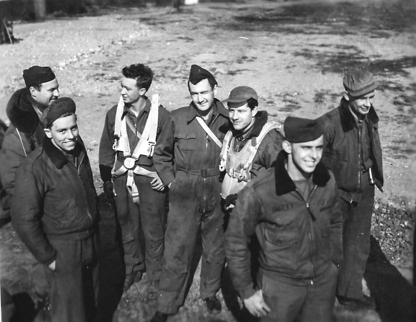 Seven of the Spirit of Martinez crew standing on gravel in flying jackets, two wearing parachute harnesses
