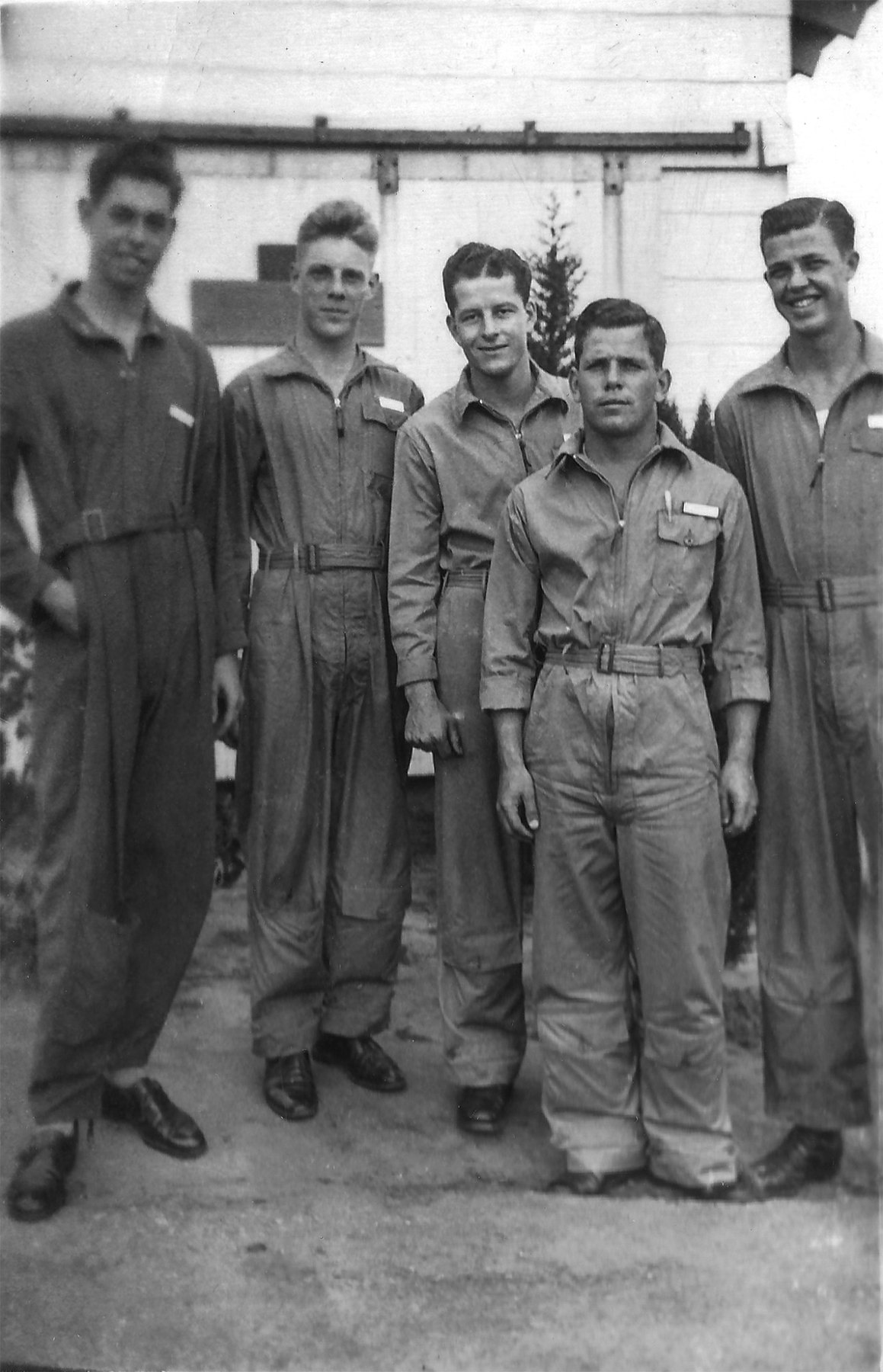 Five young men in belted flight coveralls with name tapes on the breast pockets, standing in front of a white building with a sliding door