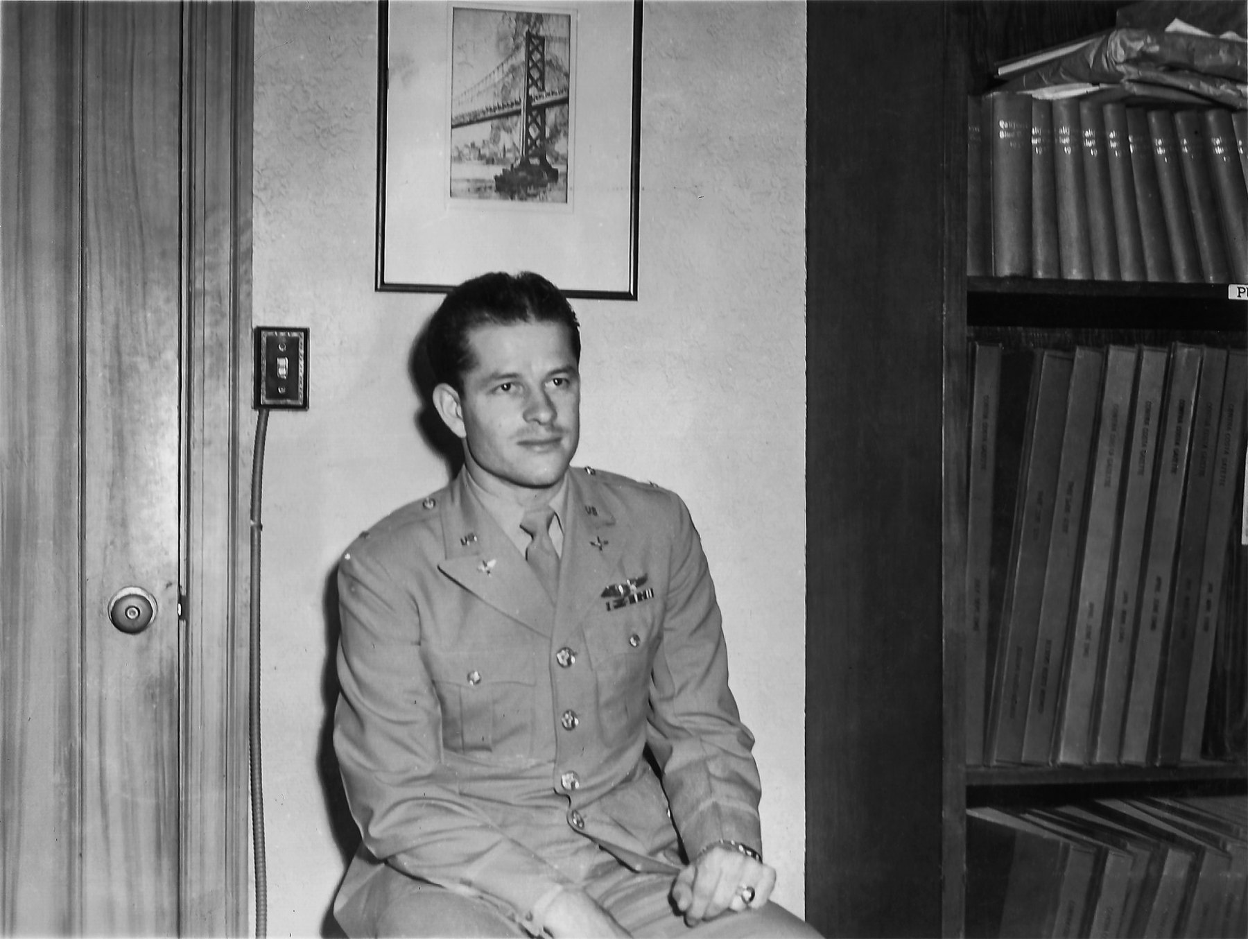 Frank Calicura in Army Air Forces service dress, seated by a door under a framed etching of the Bay Bridge, bound newspaper volumes on the shelves at right