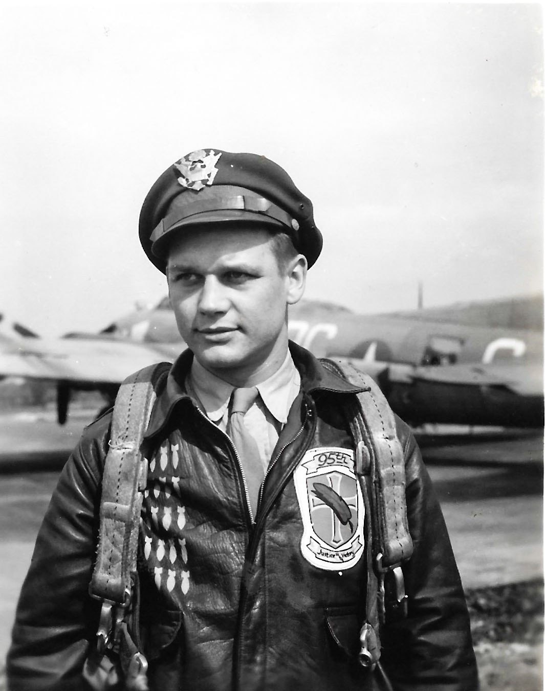 Airman in a crush cap and leather A-2 jacket with a 95th group patch and rows of painted bombs, parachute harness over his shoulders, a B-17 behind him
