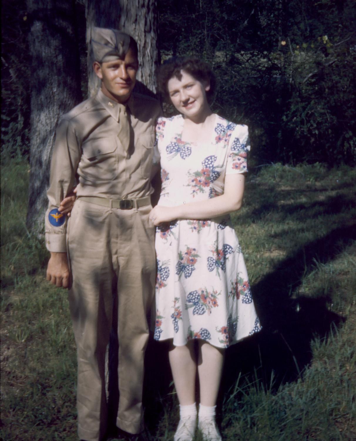 Joyce and Frank Calicura standing together under pine trees: he in khaki, she in a floral dress, Maxwell Field, 1943
