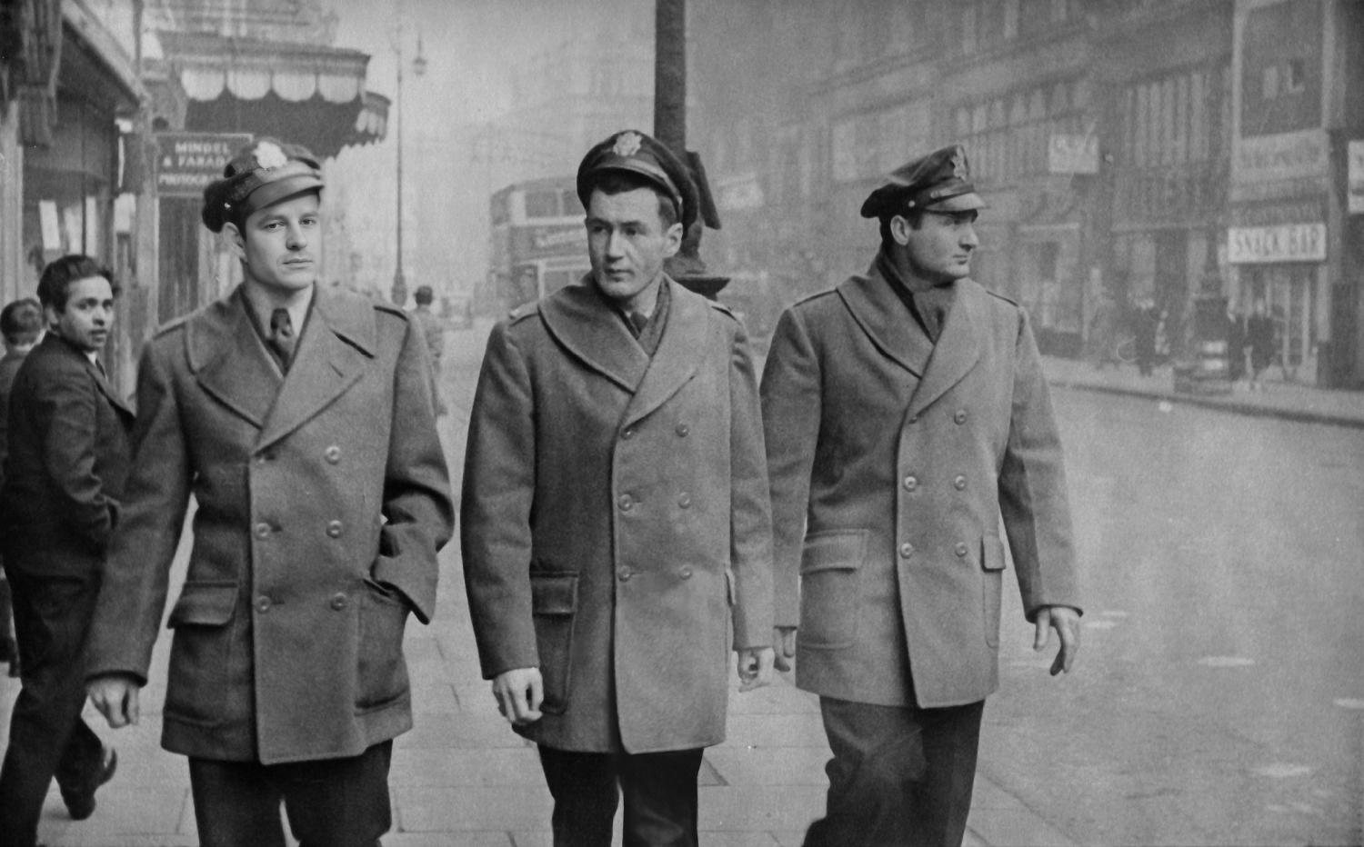 Three American officers in greatcoats walking a wet London street, a double-decker bus behind them