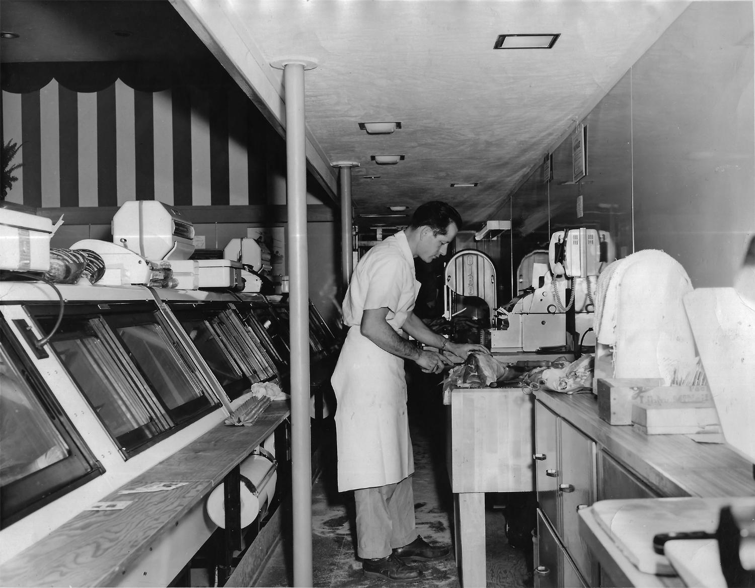 Frank Calicura in a white shirt and butcher’s apron working a cutting block in a shop lined with refrigerated cases