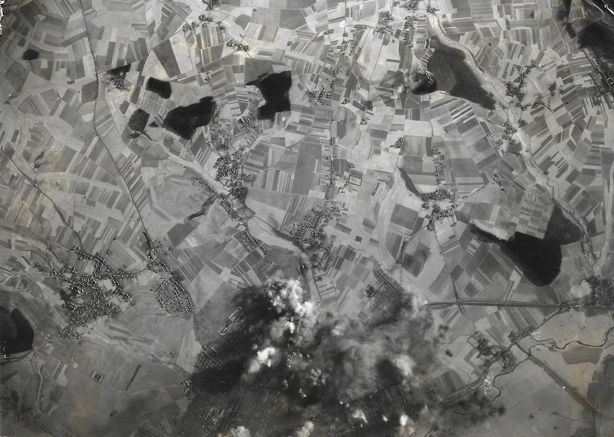 Vertical aerial photograph of strip-farmed countryside, villages and dark woods, smoke and bursts rising over a town and rail yard at the bottom of the frame